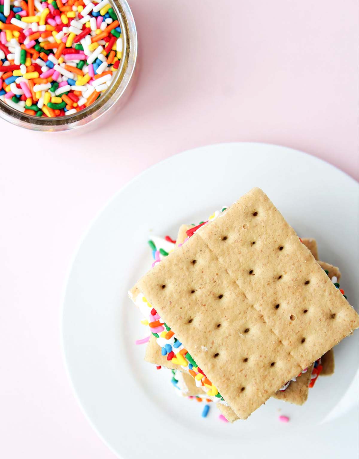 A graham cracker sandwich on a plate next to a jar of sprinkles.