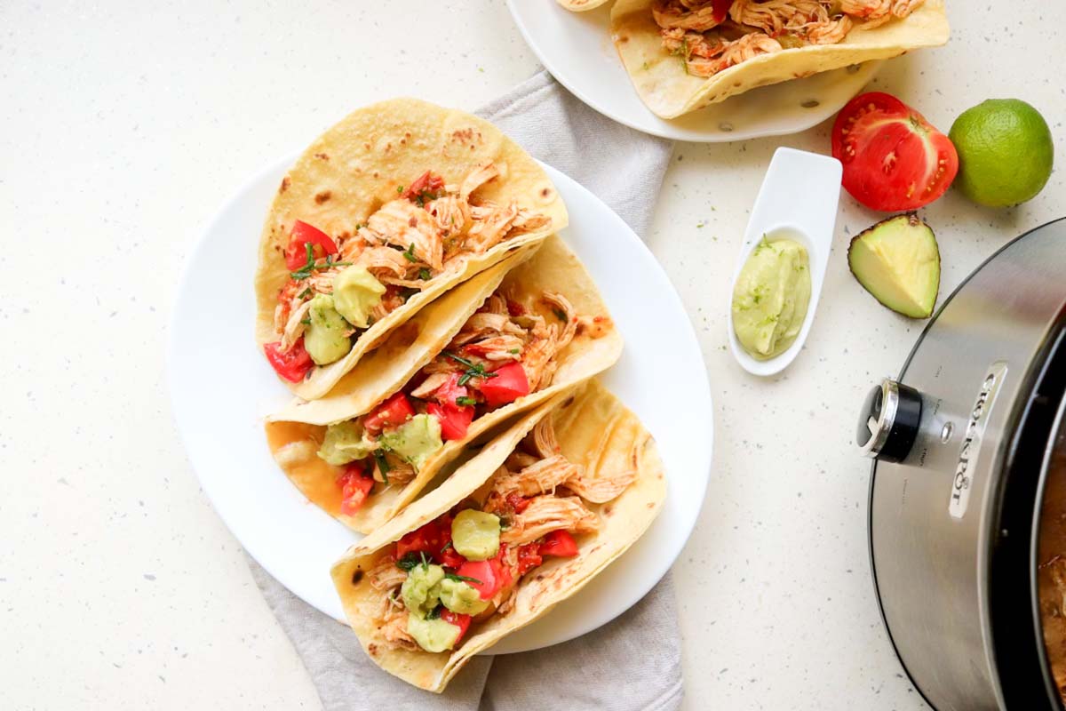 Chicken tacos on a plate next to the Crockpot.