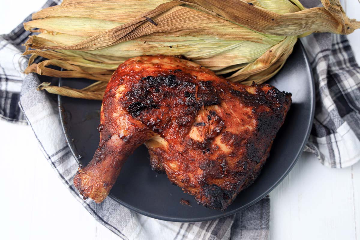 Chicken leg quarter on a black plate next to a corn husk.