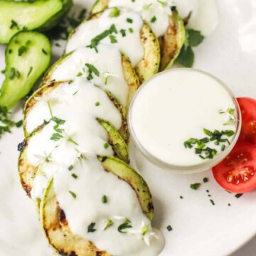 Cheese sauce in a bowl set on a white plate and drizzled over grilled zucchini.